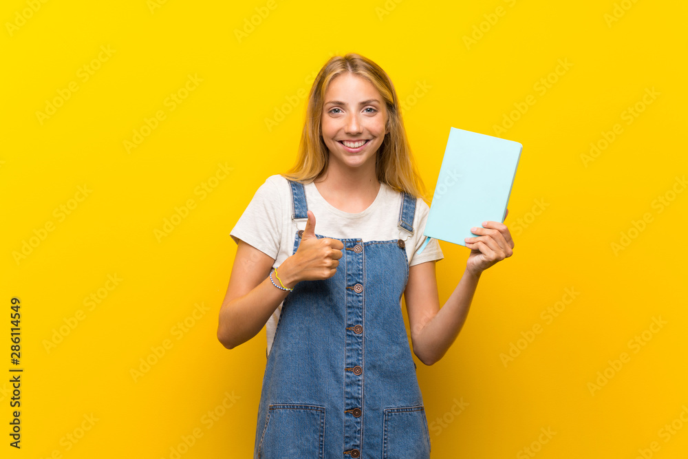Blonde young woman over isolated yellow background holding and reading a book Blonde young woman over isolated yellow background holding and reading a book
