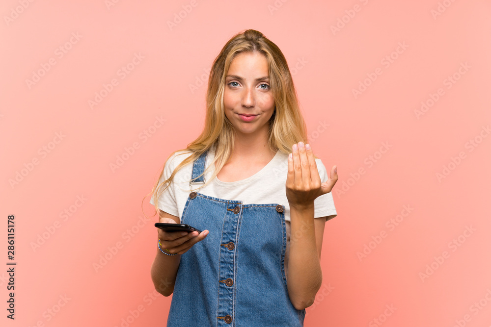 Blonde young woman with a mobile phone over isolated pink wall inviting to come with hand. Happy that you came Blonde young woman with a mobile phone over isolated pink wall inviting to come with hand. Happy that you came