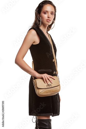 Medium full shot of a young chestnut European lady with a crossbody bag and dressed in a brown buttoned dress and dark over-the-knee boots. The bag is made of light beige leather  photo