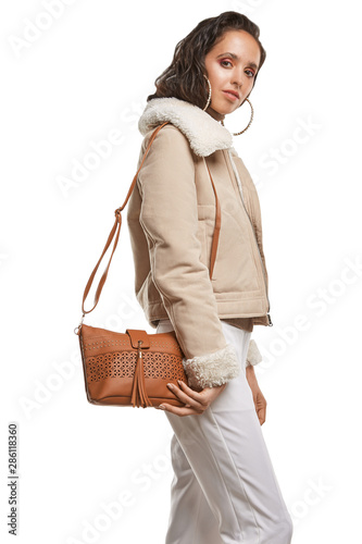 Medium full shot of a young chestnut European lady with a shoulder bag and dressed in a brown buttoned dress and dark over-the-knee boots. The bag is made of rust-coloured leather 