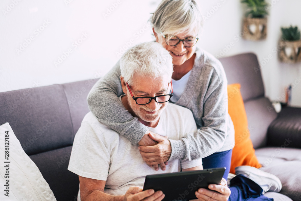 couple of seniors smiling and looking at the same tablet hugged on the sofa - indoor, at home concept - caucasians mature and retired man and woman using technology