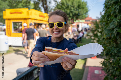 Smiling woman at an outdoor fair holds up a half-eaten footlong hot dog. Focus on the hot dog, girl intentionally blurred