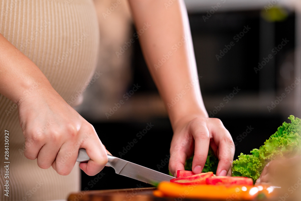Young asian pregnant woman in ketchen. Cutting tomato with large knief. Parenthood concept. Close up shot.