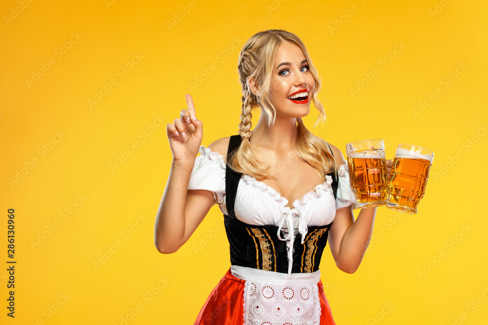 Young sexy Oktoberfest girl waitress, wearing a traditional Bavarian or german dirndl, serving big beer mugs with drink isolated on yellow background. Woman pointing to looking up. - obrazy, fototapety, plakaty 