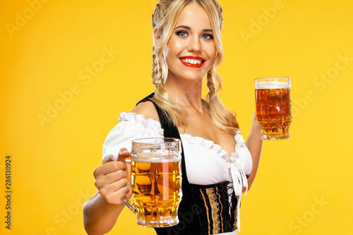 Young sexy Oktoberfest girl waitress, wearing a traditional Bavarian or german dirndl, serving two big beer mugs with drink isolated on yellow background.