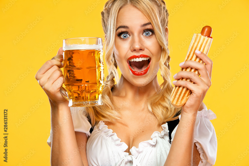 Young sexy Oktoberfest girl waitress, wearing a traditional Bavarian or german dirndl, serving big beer mugs with drink and french hot dog isolated on yellow background. - obrazy, fototapety, plakaty 