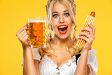Young sexy Oktoberfest girl waitress, wearing a traditional Bavarian or german dirndl, serving big beer mugs with drink and french hot dog isolated on yellow background.