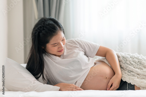 Asian pregnant woman laying in bed with open shirt. Touching her belly. Happy smile face.