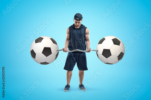 Strong muscular young man holding bar-bell with two football balls on blue background © gearstd