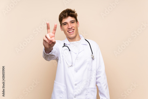 Handsome doctor young man over isolated wall smiling and showing victory sign