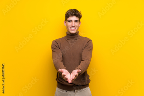 Handsome young man over isolated yellow background holding copyspace imaginary on the palm to insert an ad