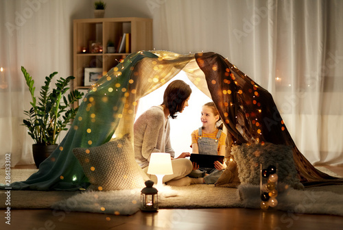 family, hygge and technology concept - happy mother and little daughter with tablet pc computer in kids tent at night at home photo