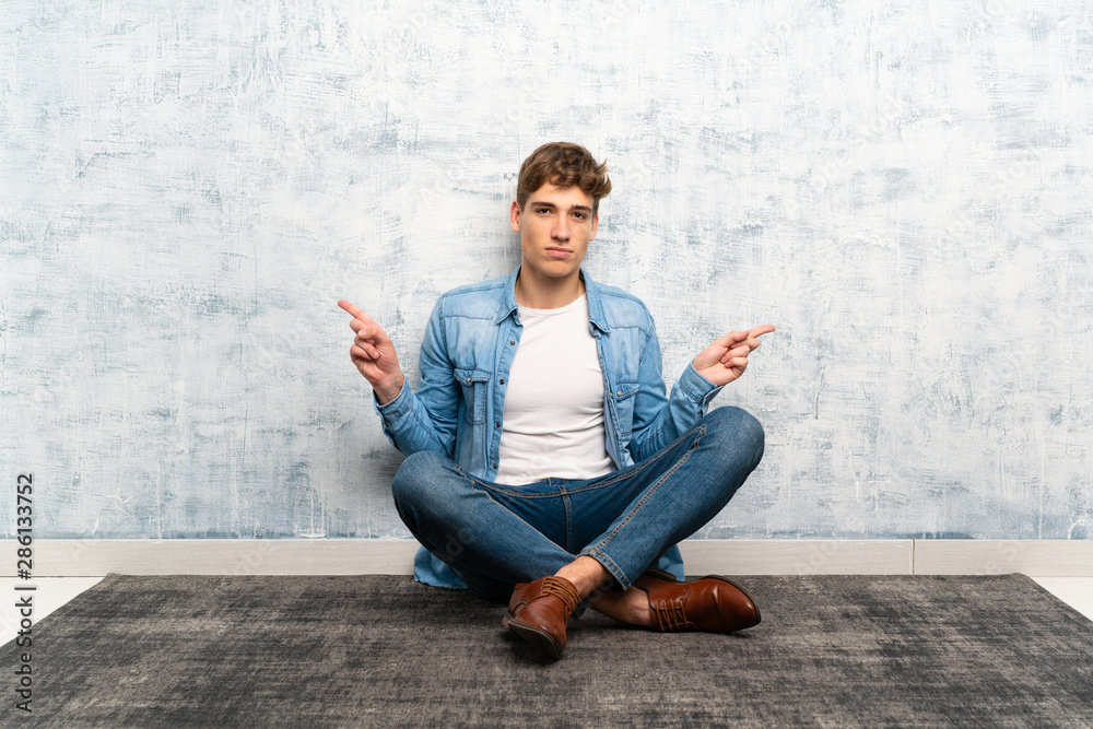 Handsome young man sitting on the floor pointing to the laterals having doubts Handsome young man sitting on the floor pointing to the laterals having doubts
