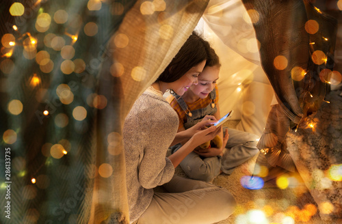 family, hygge and technology concept - happy mother and little daughter with smartphone in kids tent at night at home