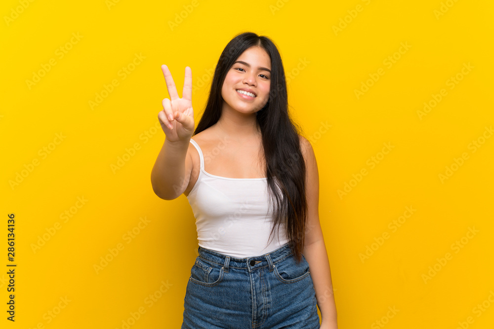 Young teenager Asian girl over isolated yellow background smiling and showing victory sign Young teenager Asian girl over isolated yellow background smiling and showing victory sign