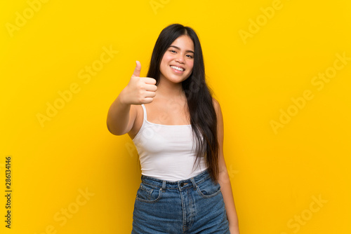 Young teenager Asian girl over isolated yellow background with thumbs up because something good has happened