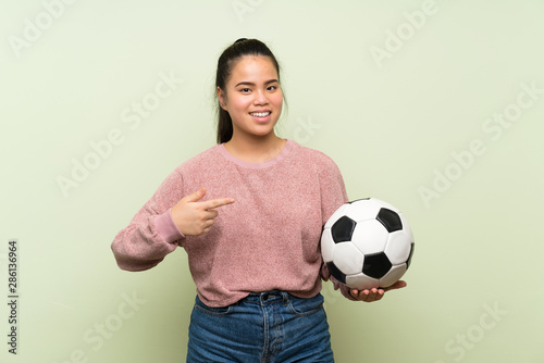 Young teenager Asian girl over isolated green background holding a soccer ball