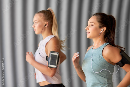 fitness, sport and healthy lifestyle concept - young women or female friends with earphones wearing armbands with smartphones and running outdoors