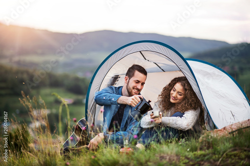 Young tourist couple travellers with tent shelter sitting in nature, drinking coffee.