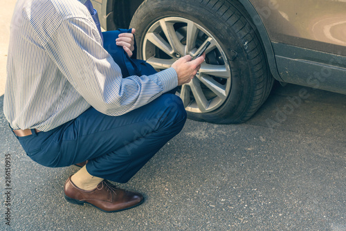 A businessman in suit crouching near his car with  punctured wheel. Man calling police using mobile phone. Street crime concept
