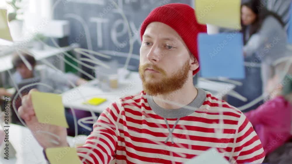 Chest-up shot of young Caucasian man with red beard and moustache in creative outfit standing in front of glass panel in studio and planning tasks for new project, and busy colleagues in background Chest-up shot of young Caucasian man with red beard and moustache in creative outfit standing in front of glass panel in studio and planning tasks for new project, and busy colleagues in background