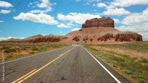 A car is driving on a road through Capitol Reef National Park. Red rock formations are seen in view. The sky is sunny with a few clouds. photo