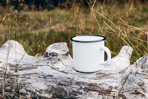 White metal mug sitting on old wood log. Blurred grass, meadow background. Outdoor tea, coffee time. Mockup of enamel cup. Lifestyle relax, trekking and camping concept. photo