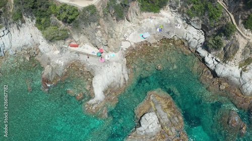 Aerial shot of Cala Trons cove with transparent blue water sparkling in sun and rocky beach near small mediterranean resort town. Drone view of beautiful seascape, Costa Brava, Catalonia, Spain photo