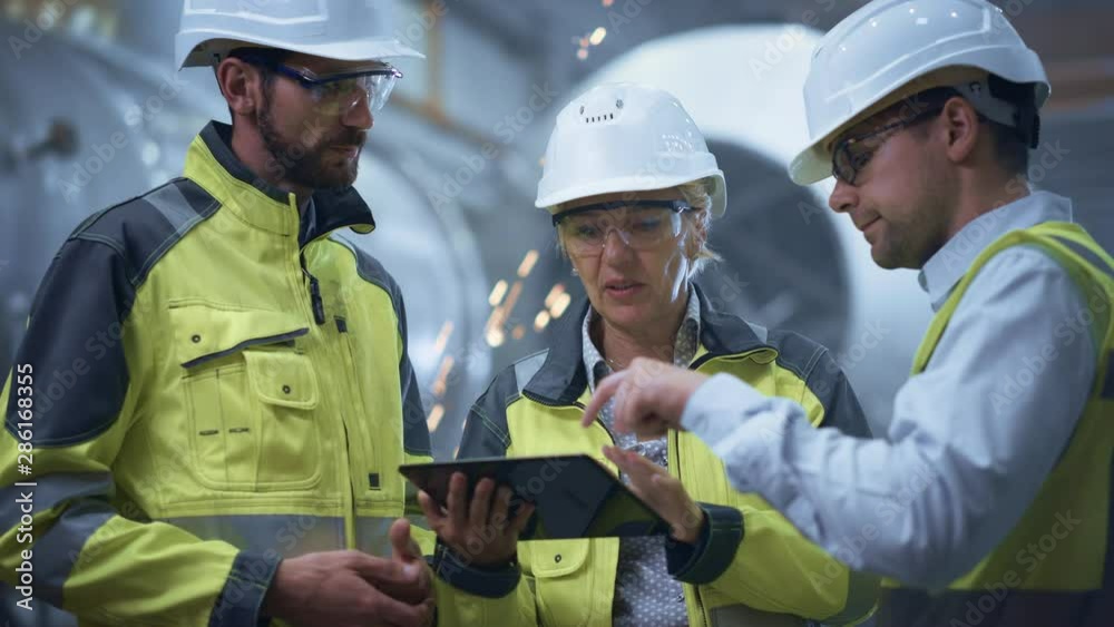 Three Heavy Industry Engineers Stand in Pipe Manufacturing Factory, Use Digital Tablet Computer, Have Discussion. Large Pipe Assembled. Design and Construction of Oil, Gas and Fuels Transport Pipeline