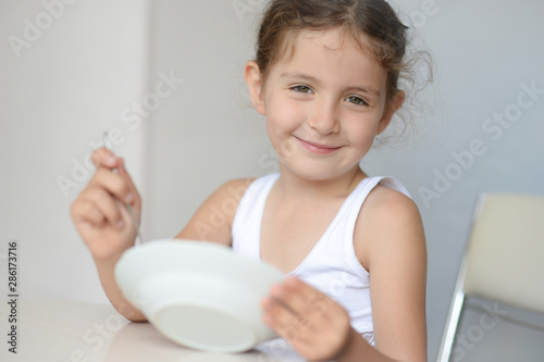 happy smiling beautiful little caucasian girl in white tank top holding spoon and plate in bright kitchen dining room