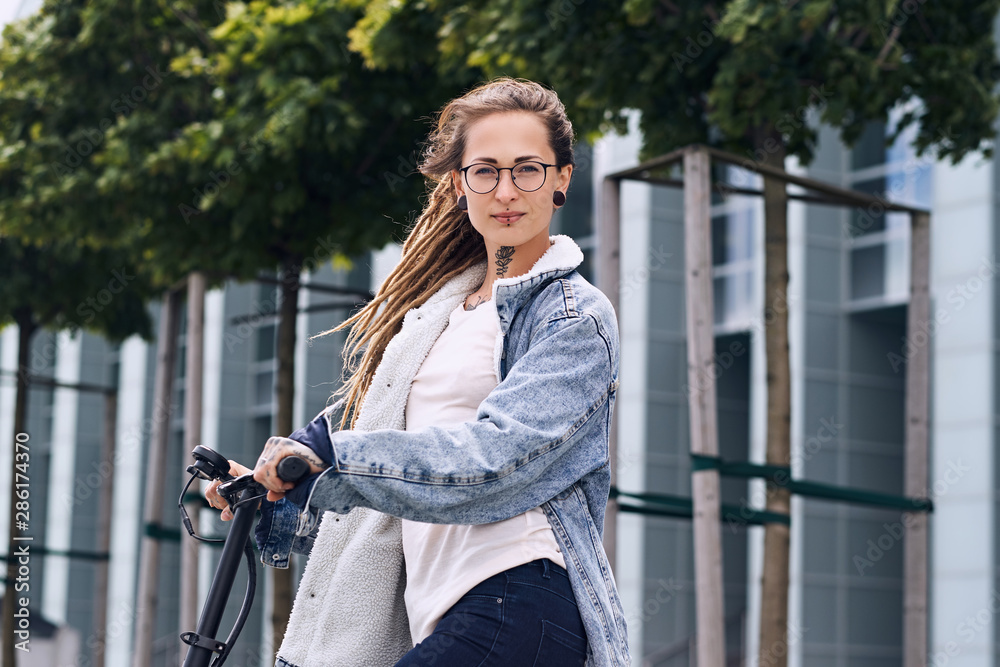 Attractive styilish girl with dreadlocks is driving electro scooter on the street near interesting building.