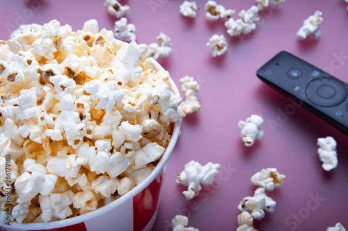 Close-up of remote control and striped bowl of popcorn on red background. The concept of watching a movie, TV, DVD. Entertainments