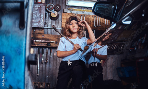 Portrait of little cheerful girl with big wrench in hands near shiny gar at auto service.