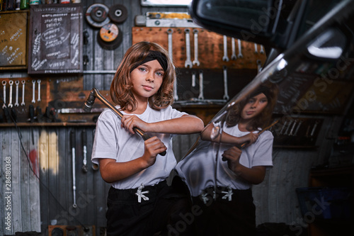 Portrait of little cheerful girl with big wrench in hands near shiny gar at auto service.