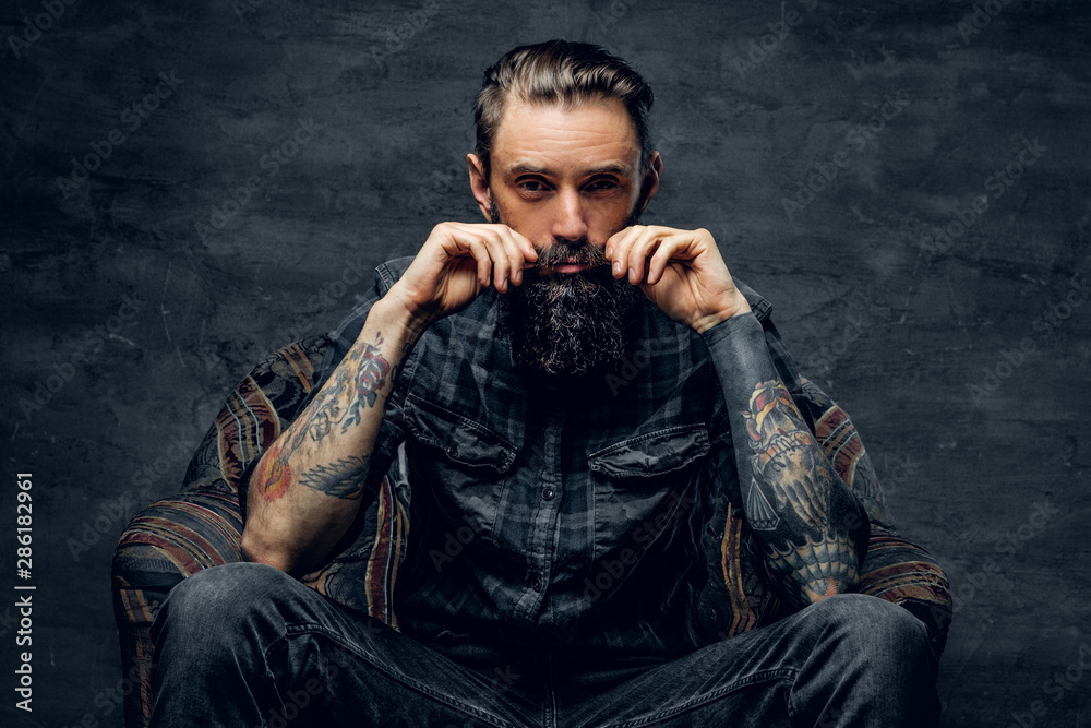 Bearded pensive man with tattooes is sitting on the armchair at dark room.