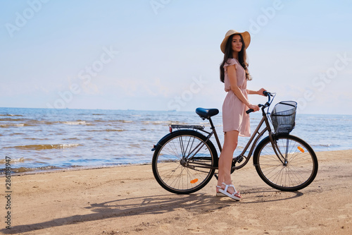 Pretty cheerful girl in hat is walking on the seaside with her bike on bright sunny day. © Fxquadro
