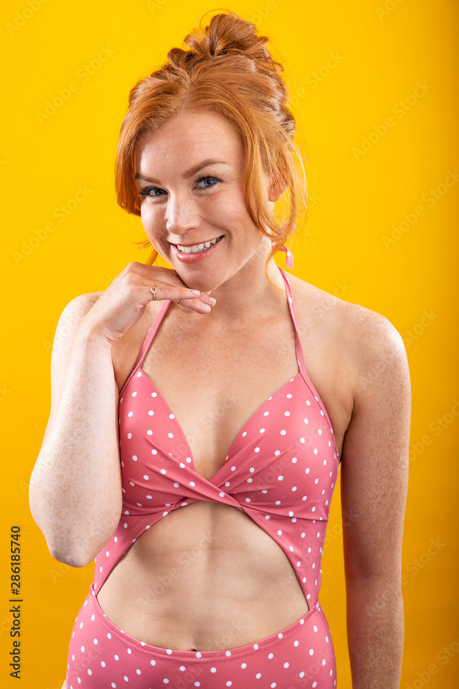 Cute girl with red hair and freckles wearing a pink polka dot bikini with a yellow background Adobe Stock
