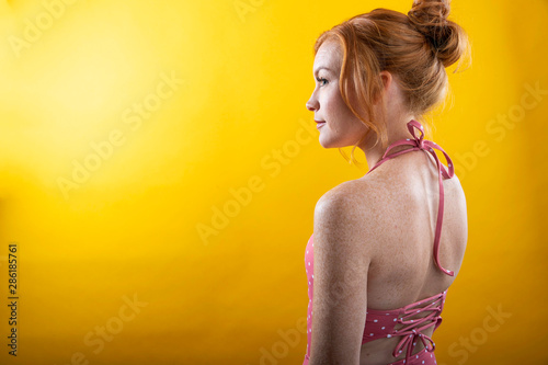 Cute girl with red hair and freckles wearing a pink polka dot bikini with a yellow background