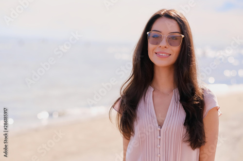 Portrait of beautiful smiling girl in sunglasses and pink dress at calm seaside.