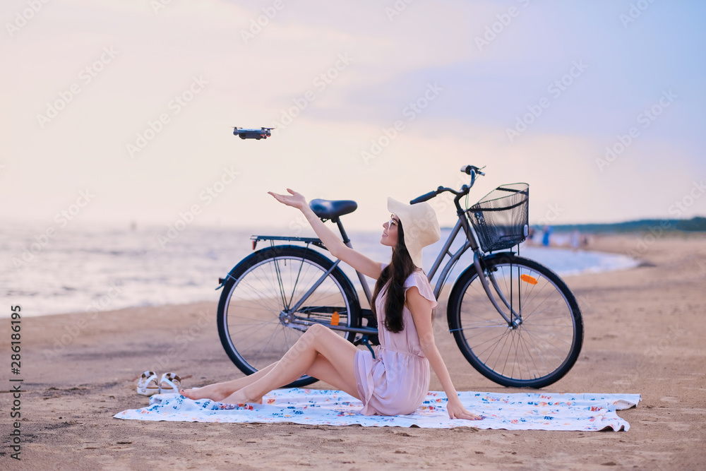 Obraz premium Pretty happy woman is trying to catch a drone while chilling on the beach next to her bike.