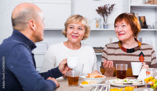 Smiling aged friends are sitting at table with tea and talking about life