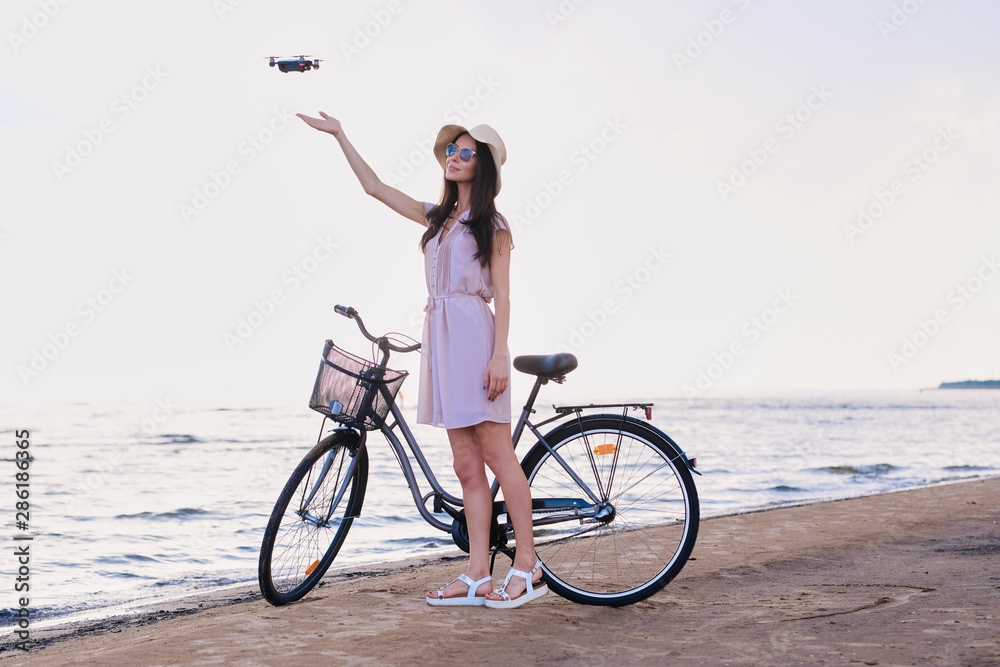 Fototapeta premium Pretty happy woman is trying to catch a drone while chilling on the beach next to her bike.