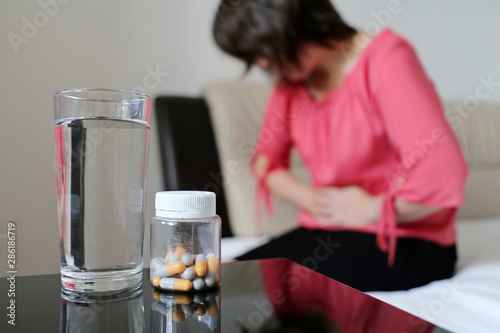 Stomach ache, bottle of pills and water glass on background of woman suffering from abdominal pain. Girl sitting on bed clutching her abdomen, concept of indigestion, menstruation photo