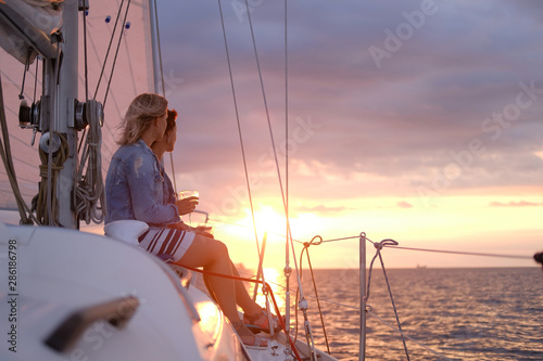 Couple of ladies are enjoying their trip on sailing boat while drinking wine and watching beautiful sunset.