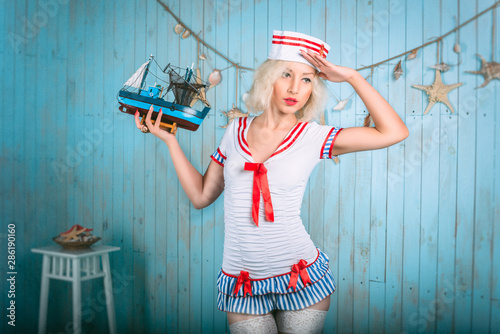 Sexy young blonde woman salutes in sailor costume with red stripes and holds ship in hand over blue background with shells. Sea theme