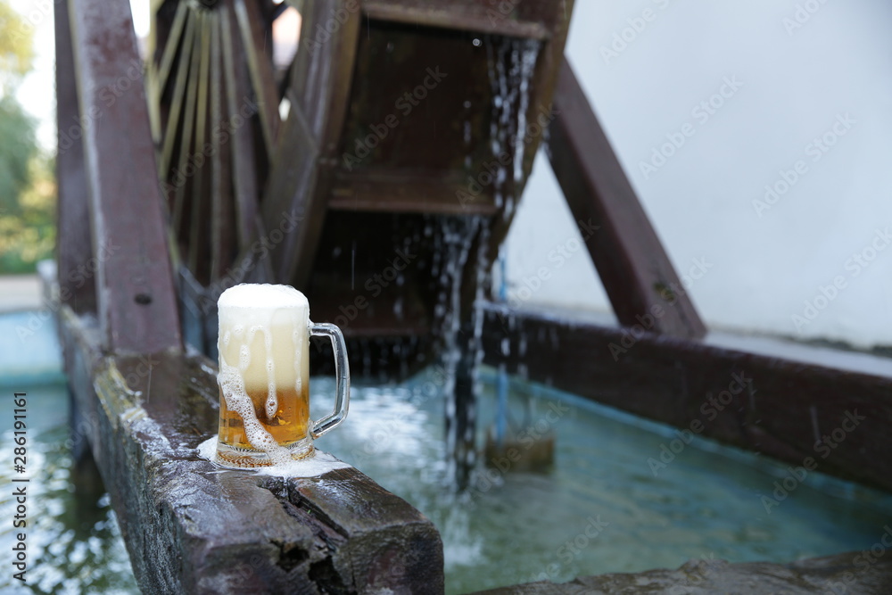 Glass of beer with foam on a natural wooden board on a background of a wooden mill wheel with falling water. Pattern, banner, background.