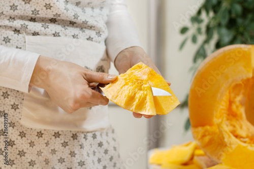 Kobiece dłonie trzymają kawałek dyni i obierają go ze skóry za pomocą noża kuchennego. Połowa świeżej dużej pomarańczowej dyni leży na kuchennym blacie.