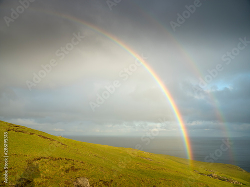 rainbows in the sky with clouds above green hill with sea view