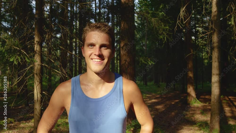 PORTRAIT, SLOW MOTION, CLOSE UP: Happy Caucasian male jogger smiles after finishing a fun trail run in the forest on a sunny summer day. Sweaty man proudly stands in front of the woods after a jog. PORTRAIT, SLOW MOTION, CLOSE UP: Happy Caucasian male jogger smiles after finishing a fun trail run in the forest on a sunny summer day. Sweaty man proudly stands in front of the woods after a jog.