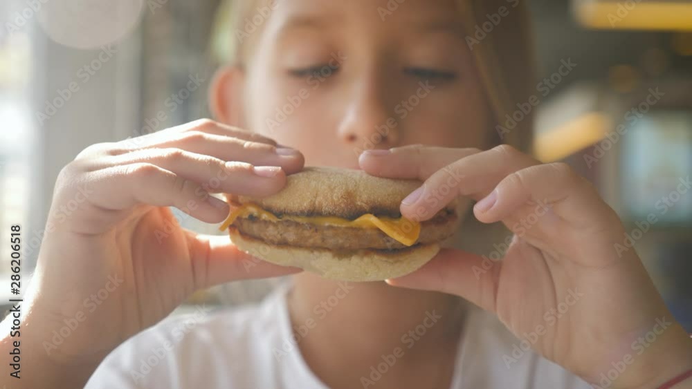 Beautiful happy hungry baby girl eating hamburger. Fast food concept. Tasty unhealthy burger sandwich in hands.